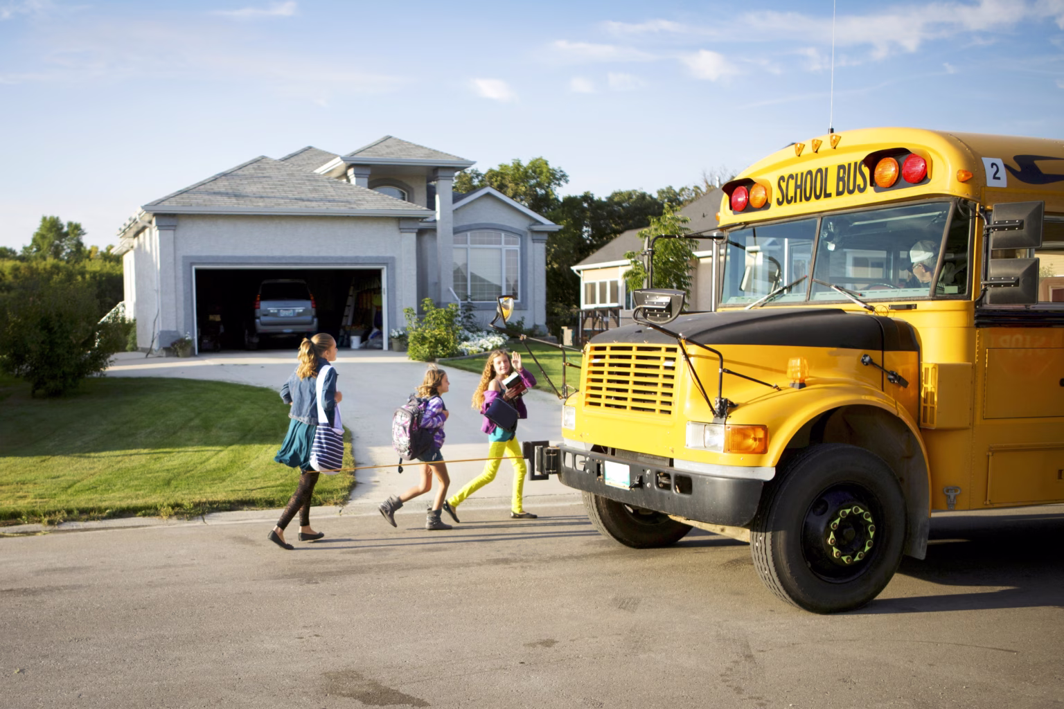 Three children with backpacks walking to a yellow school bus on a residential street