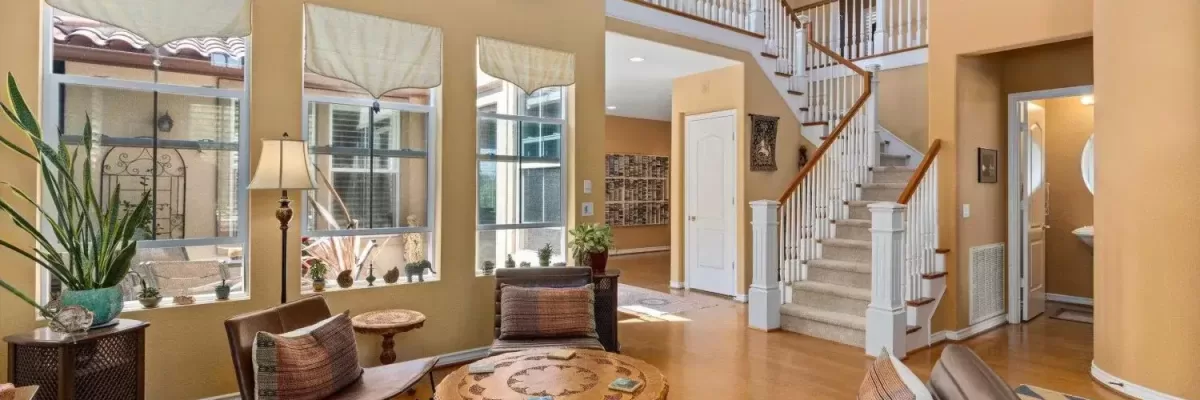 Interior view of the soaring double-height living room at 2198 Vista La Nisa in Carlsbad, CA. Features warm hardwood flooring, tall windows overlooking the courtyard with accent stained-glass cutouts above, and a grand open staircase leading to the upper-level banister and gallery hallway.